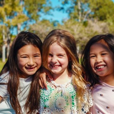 three smiling girls with long hair in the sunshine there is grass and trees behind them